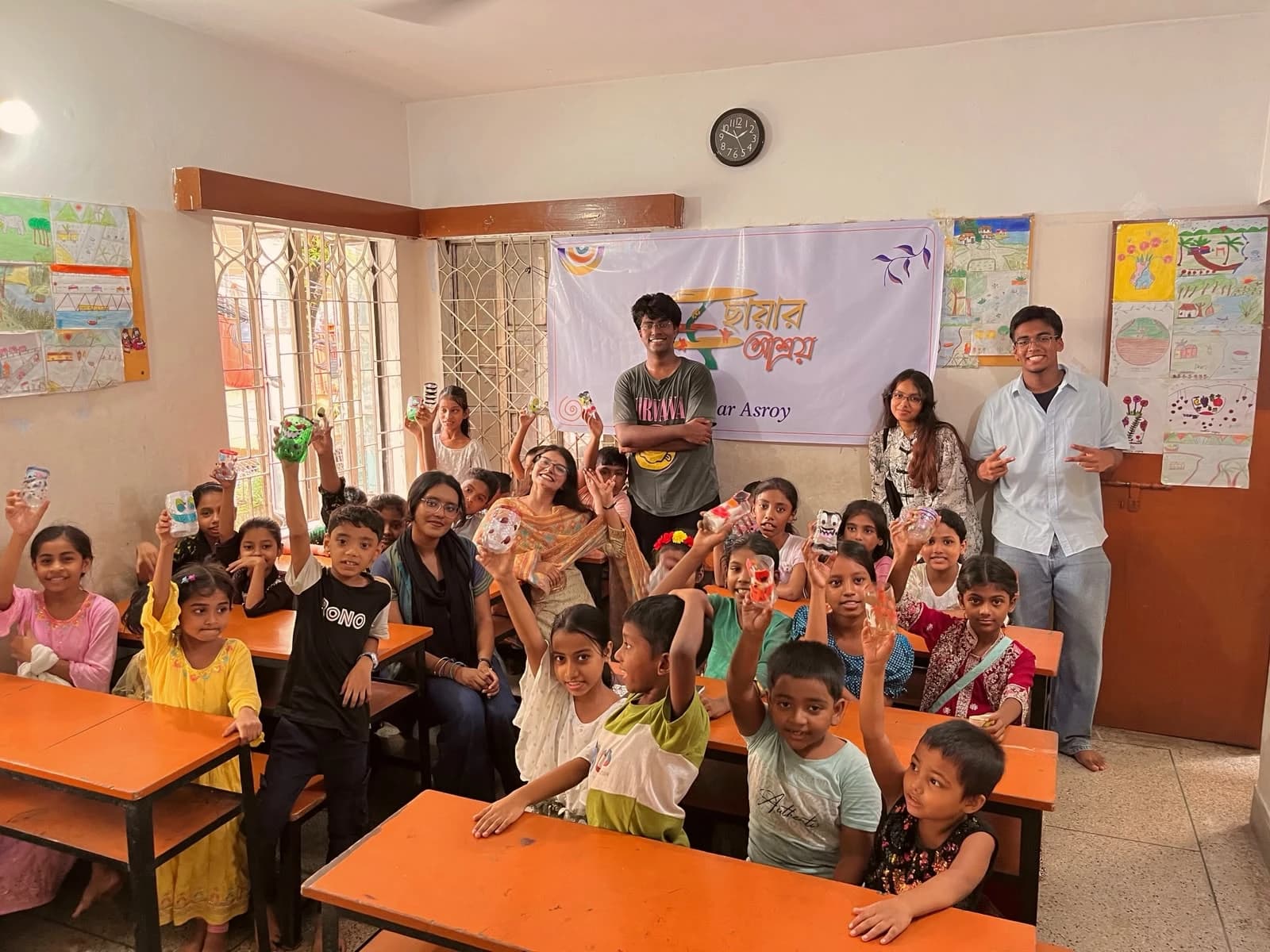 Volunteers and children holding up their finished bottle pen holders in front of the Chayar Asroy banner